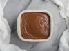 Fotografia vista de cima de um recipiente quadrado com a sobremesa natalina, finalizada com ganache de chocolate sobre uma mesa de mármore com pano branco em volta.