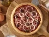 Fotografia em tons de marrom em uma bancada de madeira, uma tábua de madeira redonda com a pizza de calabresa em cima dela. Ao lado, cebola, potinho de azeite e cortador de pizza.