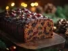Fotografia de um bolo de frutas natalino, com cobertura de chocolate e decorado com amêndoas e uvas-passas, cercado por pinhas e ramos de pinheiro, transmitindo um clima festivo.
