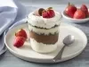 Imagem de um copo com a receita mostrando as camadas de creme, decorado com morango e um bombom. Ao fundo, um potinho com morangos.