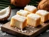Fotografia de pedaços fofinhos de bolo de coco dispostos em uma tábua de madeira, com cobertura de coco ralado e uma crosta dourada.
