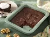 Fotografia em tons de verde em uma bancada de madeira verde, um recipiente verde quadrado com o brownie de chocolate com nozes e um pano colorido ao lado. Ao fundo, um prato redondo bege com uma fatia do brownie.