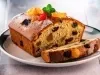 Fotografia de uma fatia de bolo de frutas em um prato, com uma crosta dourada e interior úmido, repleto de frutas secas. O bolo é decorado com açúcar de confeiteiro, folhas de hortelã e frutas cristalizadas.