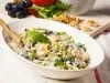 Fotografia de uma salada em uma tigela oval branca, com maçãs, melão e uvas pretas em um molho cremoso, decorada com nozes e salsa, acompanhada de um garfo, em um ambiente rústico com ingredientes frescos ao fundo.