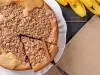 Fotografia vista de cima de uma cuca de banana com farofa sobre um prato de vidro branco raso, apoiado em uma mesa de madeira preta com um pano marrom de tom claro.