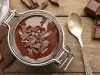 Fotografia vista de cima de um recipiente de vidro, tipo um copo, que fecha sobre uma mesa de madeira. Dentro tem a sobremesa, finalizada com ganache e raspas de chocolate, e ao redor tem uma colher pequena e alguns pedaços de chocolate.