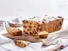 Fotografia de um bolo de frutas recém-assado em uma tábua de madeira, com fatias expostas que mostram sua textura densa e recheio colorido de frutas secas, em uma cozinha iluminada.