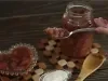 Fotografia em tons escuros de uma mesa de madeira escura, sobre ela tem dois apoios quadriculados de madeira. Apoiado neles tem um pote de vidro com doce de banana, um pote de vidro pequeno em formato de coração e uma mão segurando o doce na colher.
