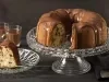 Fotografia em tons de marrom e preto de uma bancada preta vista de frente. Contém um prato redondo transparente com uma fatia de bolo com calda de chocolate por cima, um recipiente transparente para servir o bolo com o bolo sem uma fatia e calda por cima.