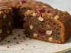 Fotografia de um bolo de frutas, redondo e bem assado, com uma fatia cortada que revela um interior denso, repleto de nozes e frutas cristalizadas. O bolo é decorado com cerejas vermelhas.