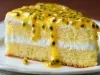 Fotografia de um bolo gelado de maracujá, apresentado em um prato floral. O bolo é de duas camadas, coberto com um molho amarelo brilhante, decorado com folhas de hortelã e frutas escuras.