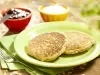 Fotografia em tons de marrom e verde de uma bancada de madeira com um paninho verde, sobre ele um prato redondo verde com dois pães de frigideira. Ao fundo dois recipientes redondos vermelho e amarelo com geleia e patê e uma faca.