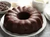 Bolo de chocolate em forma de bundt, com cor marrom escura e superfície brilhante, servido em um prato branco. Ao fundo, um jarro de cerâmica branca e pedaços de chocolate.
