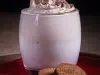 Foto da receita de cappuccino super cremoso servida em um copo de vidro sobre uma louça vermelha em cima de uma mesa de madeira