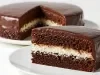 Fotografia de um bolo de chocolate redondo, coberto com ganache suave. Uma fatia revela camadas de chocolate fofinho e um recheio branco de coco.