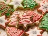 Em cima de uma mesa, prato com biscoitos em formato de estrelas, sinos e Árvore de Natal, decorados com glacê branco, verde e vermelho e confeitos coloridos de bolinha.