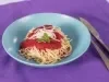 Fotografia em tons claros de um prato de vidro fundo azul sobre um pano roxo. Ao lado tem talheres juntos, como o garfo e a faca, e dentro do prato tem o mcarrão tradicional com o molho de tomate e ascenda.