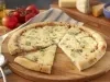 Imagem de uma pizza redonda de 4 queijos com uma fatia deslocada. A fundo uma tábua de queijos e tomates.