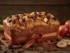 Fotografia de um bolo de frutas decorado com nozes e frutas secas, amarrado com uma fita marrom. Ao lado, maçãs vermelhas e fatias de maçã complementam a apresentação.