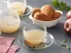 Imagem de uma xícara de chá de maçã, com um pedaço de canela no pires ambientado com maçãs e um pote de madeleines