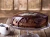 Fotografia de um bolo de chocolate em um suporte de vidro, coberto com ganache escura que escorre pelas laterais. Ao lado, um pequeno jarro de cerâmica com ganache, em um cenário acolhedor de madeira.