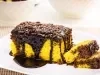 Pedaço de bolo de cenoura com cobertura de chocolate e granulado visto de outro ângulo. A calda escorre pelas laterais e se acumula no prato, criando um visual apetitoso. As fatias ao lado ainda aparecem, com tigelas brancas e um copo de suco.