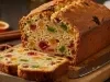 Fotografia de um bolo de frutas recém-assado, exibido em uma tábua de madeira. O bolo, cortado, mostra um interior denso e úmido, repleto de frutas cristalizadas.