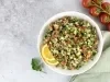 Fotografia em tons claros vista de cima de um recipiente fundo, branco, com uam receita de tabule. O recipiente está ao lado de alguns tomates, um pano verde claro e folhas de salsinha, sobre uma mesa de cimento queimado e tom claro.
