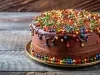 Fotografia de um bolo de chocolate redondo, coberto com uma suave cobertura de chocolate e decorado com coloridos confeitos. O bolo está sobre um prato dourado, contrastando com a superfície de madeira rústica.