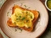 Fotografia em tom claro de uma torrada com creme de queijo parmesão e cebolas, salpicada com salsinha. A torrada está sobre um prato de tom rosa claro.