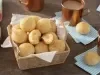 Fotografia em tons de azul em uma bancada de madeira escura, guardanapos de pano azul claro, uma cesta de vime com vários pães de queijo, xícaras cobre com café com leite dentro.