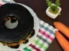 Bolo de cenoura com cobertura de chocolate, acompanhado de duas cenouras frescas e uma pequena planta decorativa. A composição é realçada por um pano xadrez colorido, tudo sobre uma superfície de madeira escura, transmitindo um ambiente acolhedor.