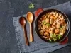 Imagem vista de cima de uma panela com cabo vermelho com a receita de cassoulet de feijão branco com linguiça e folhas de coentro decorando. Ao lado, duas colheres de pau e um pano azul escuro.