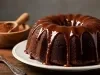 Um bolo de chocolate Bundt, rico e úmido, é apresentado em um prato cerâmico branco. Coberto com um brilho de chocolate cremosa, o bolo exibe uma textura macia e escura.