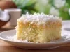 Fotografia de um bolo cortado em quadrado em um prato branco, com interior amarelo fofinho, úmido e cobertura de coco. O fundo natural com folhas verdes e um coco cortado.