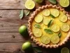 Fotografia vista de cima de uma torta de limão sobre uma mesa de madeira escura. A torta de limão está decorada com fatias do fruto, gelatina e algumas folhas.