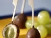Fotografia em tons de amarelo, chocolate e azul, mostrando sobre bandeja amarela três espetinhos com chocolate, sendo um aberto e recheado com uva, algumas uvas verdes, tudo sobre guardanapo e bancada em tons de azul.