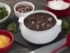 Fotografia em tons de vermelho em uma bancada de madeira escura, uma tábua preta com uma panela branca com a feijoada dentro dela. Ao lado, vários potinhos com laranja, couve, arroz e farofa.