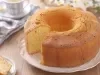 Um bolo redondo e dourado com furo no meio, servido sobre um prato branco com bordas. Uma fatia está cortada e colocada ao lado. O fundo tem uma xícara listrada, um guardanapo dobrado e flores brancas, criando um clima leve e acolhedor.