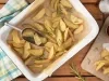 Imagem vista de cima de uma travessa retangular branca com batatas fritas rústicas, ramos de alecrim e um pegador. Ao lado, uma tábua com um saquinho de batatas, um potinho de ketchup e um potinho de sal.