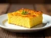 Fotografia de uma fatia de bolo de fubá com erva-doce, apresentada em um prato cerâmico branco. A crosta dourada e o interior amarelo vibrante sugerem uma textura fofa, decorada com ervas verdes frescas.