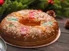 Fotografia de um bolo decorado com frutas cristalizadas vermelhas e verdes, amêndoas fatiadas e açúcar. Apresentado em um prato prateado sobre uma superfície de madeira escura, rodeado por ramos de pinheiro e enfeites natalinos.