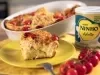 Fotografia em tons de amarelo em uma bancada de madeira, um pano amarelo e um prato amarelo redondo ao centro com um pedaço de torta de frango em cima. Ao fundo um  recipiente com a torta inteira e a lata de Ninho Adulto ao lado.