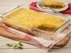 Imagem de uma travessa retangular transparente com a receita de escondidinho com carne desfiada sem um pedaço, mostrando a camada de carne e a camada de purê. Ao fundo, um prato branco com o pedaço e um garfo.