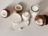Fotografia vista de cima de duas garrafas decorativas de vidro, uma jarra branca e um copo grande e largo. Todos esses recipientes têm bebida vegetal de coco. Ao lado tem cascas, pedaços e polpa do coco pela metade sobre uma mesa cinza clara.