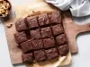 Fotografia vista de cima de uma tábua de madeira sobre uma mesa branca. Apoiado na tábua tem vários pedaços de brownie cortados e apoiados em papel manteiga.
