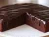 Em cima de uma mesa, prato redondo com bolo de chocolate redondo e cobertura de chocolate cremoso.