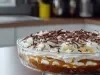 Em cima de uma mesa, refratário com biscoitos crocantes, doce de leite e chantilly, decorado com bananas em rodelas e raspas de chocolate.