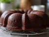 Fotografia de um bolo de chocolate em uma grade de resfriamento, com cor marrom escuro e superfície brilhante. O fundo desfocado mostra uma cozinha aconchegante, criando uma atmosfera convidativa.