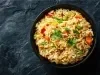 Em um prato há arroz soltinho com uma mistura de legumes, como cenoura, ervilhas e pimentões, decorado com folhas de salsinha.